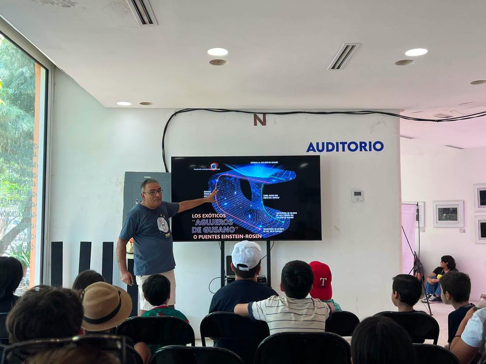 Un instructor de SAMAZ señala la pantalla del auditorio, que muestra la lámina «Los exóticos agujeros de gusano o puentes Einstein-Rosen», frente a niñas y niños sentados.