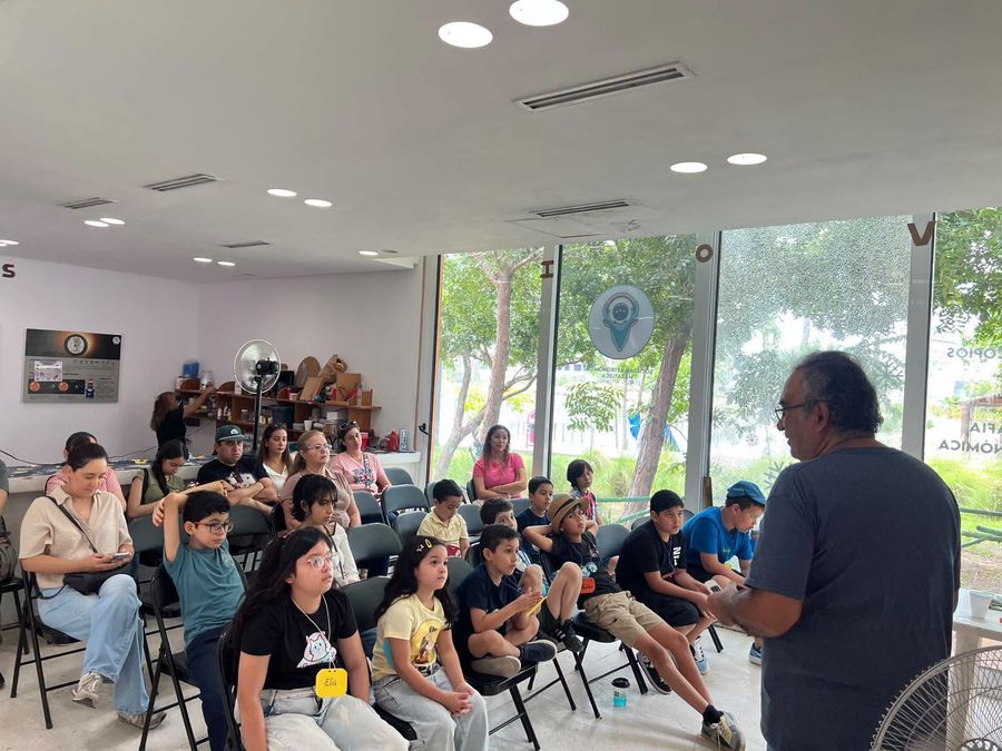 Niñas y niños sentados escuchando la charla del domingo en la sede de SAMAZ, con sus papás atrás.