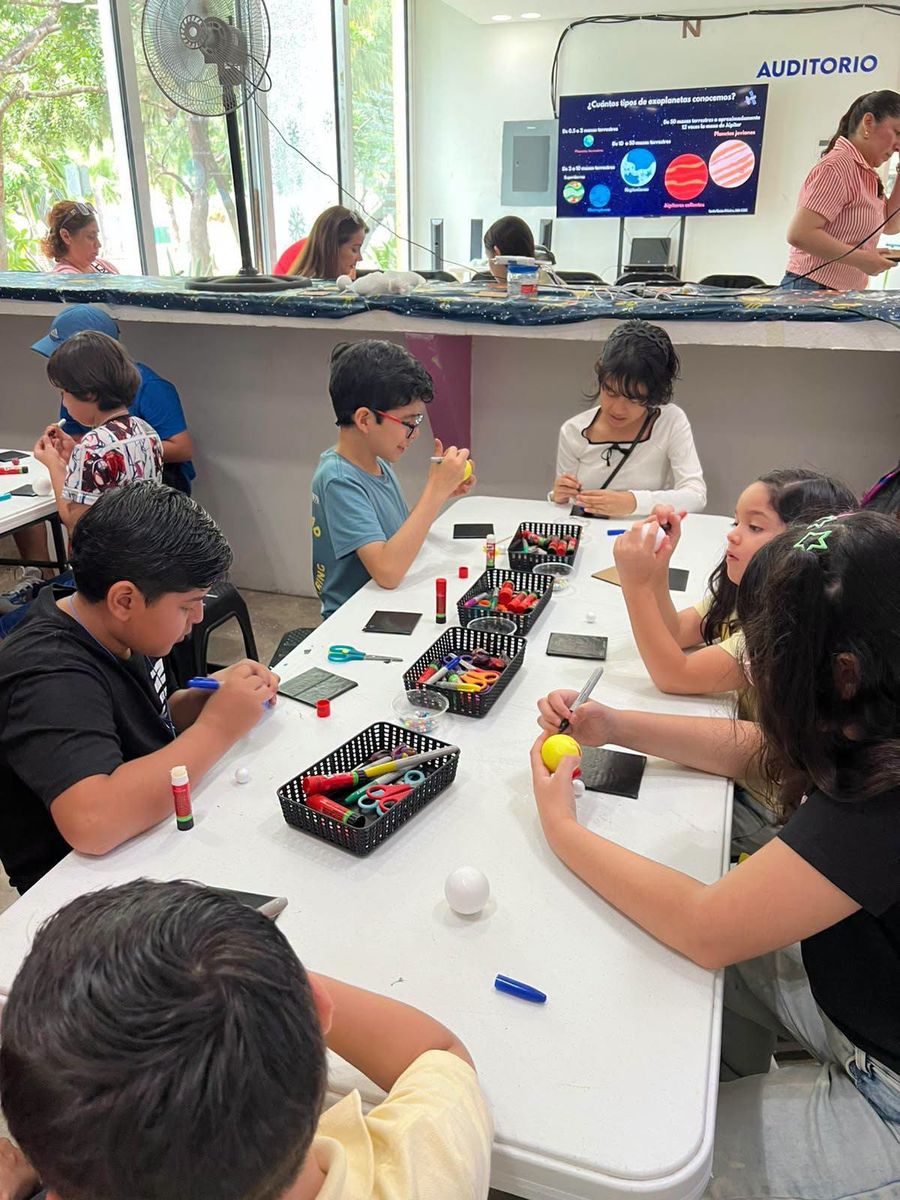 Niños pintando esferas de unicel en una mesa; al fondo, la pantalla del auditorio con los tipos de exoplanetas.