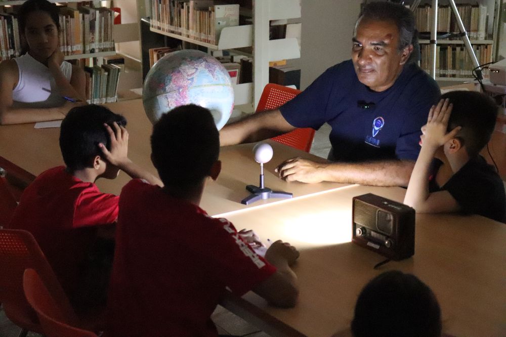Daniel Hernández, coordinador del club, hace una demostración de eclipse con un globo terráqueo y una esfera iluminada mientras las niñas y los niños observan sentados a la mesa.